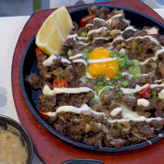 Chopped beef sisig topped with a raw egg yolk, mayonnaise drizzle, spring onions and red chillies, with a lemon wedge, on a black cast-iron sizzling plate set on a dark red wooden board at Pinoy Tambayan