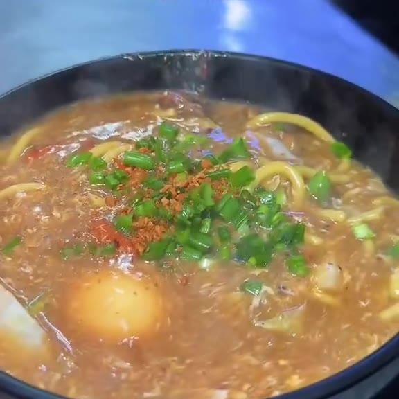 Close-up of a black bowl of lomi with thick brown gravy, yellow egg noodles, chopped spring onions, toasted garlic bits and a soft-cooked egg at Pinoy Tambayan
