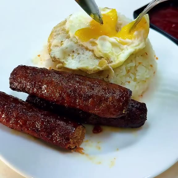 Two browned longganisa sausages in front of a sunny-side-up egg with a broken runny yolk over garlic fried rice on a white plate at Pinoy Tambayan