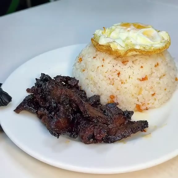 Fried egg resting on a mound of garlic fried rice beside dark caramelised beef tapa strips on a round white plate at Pinoy Tambayan