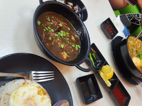 Overhead view of a shared table with crispy chicken skin, a dark braised stew topped with spring onions, and garlic rice with a fried egg.