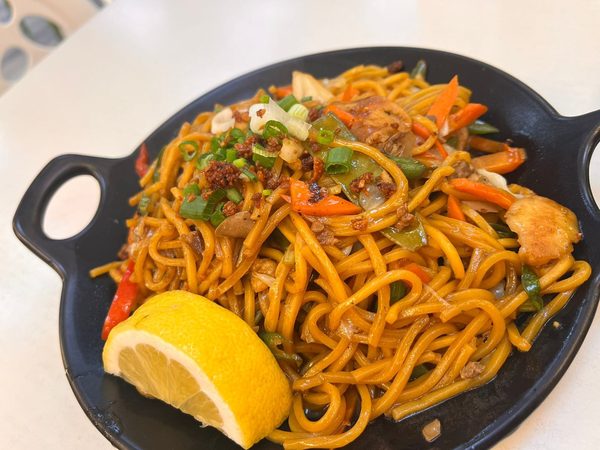 Pancit canton noodles on a black serving plate with carrot, green pepper, cabbage and spring onion, topped with crisp fried garlic and a lemon wedge.