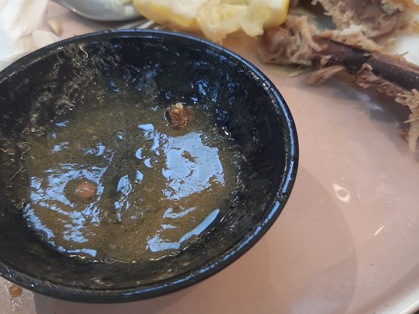 An almost empty black bowl of dark sauce beside picked-over bones, boiled cabbage leaves and squeezed lemon on a pale plate.