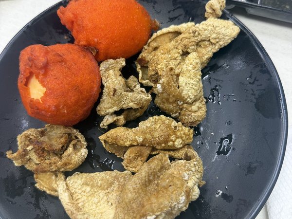 Black plate of golden crispy chicken skin with two orange-battered kwek kwek eggs, beside a tray of three dipping sauces.