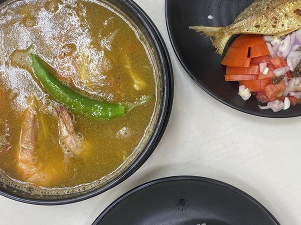 Overhead spread of prawn sinigang with a green chilli, two whole fried fish with tomato and onion salad, glazed sausages and steamed rice.