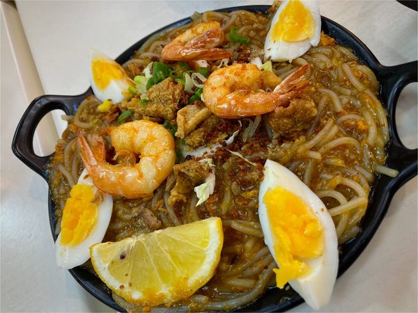 Pancit Malabon on a black cast iron platter, topped with prawns, quartered boiled eggs, crispy chicken skin, spring onion, toasted garlic and a lemon wedge.