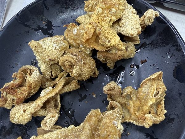 Curled pieces of crispy golden chicken skin on a black plate, with dark and red dipping sauces alongside.