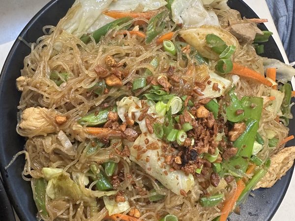 Pancit bihon in a black handled dish, thin noodles tossed with cabbage, carrot, mangetout, chicken, spring onion and crisp fried garlic.