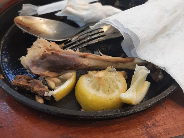 A finished sizzling plate with a bone picked almost clean, roasted garlic cloves, squeezed lemon and used cutlery on a wooden board.