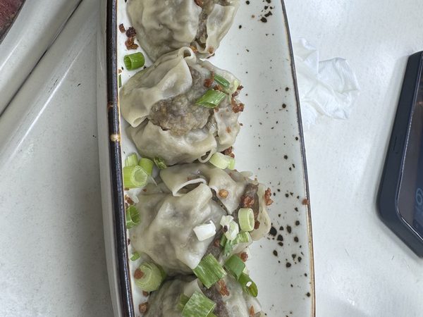 Four steamed siomai dumplings topped with spring onion and crisp fried garlic on a long white plate, with a phone face-up at the table edge.