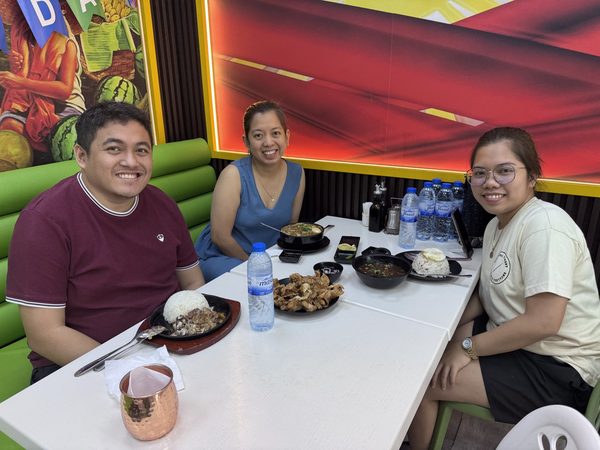 Three adult diners smile at the camera around sizzling sisig, rice, a plate of crispy chicken skin, soup bowls and garlic rice with a fried egg.