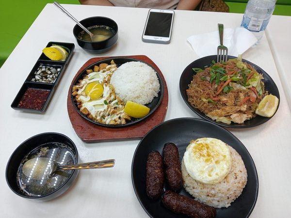 Overhead spread of sizzling sisig with egg and rice, pancit bihon, browned sausages with garlic rice and fried egg, soup bowls and condiments.